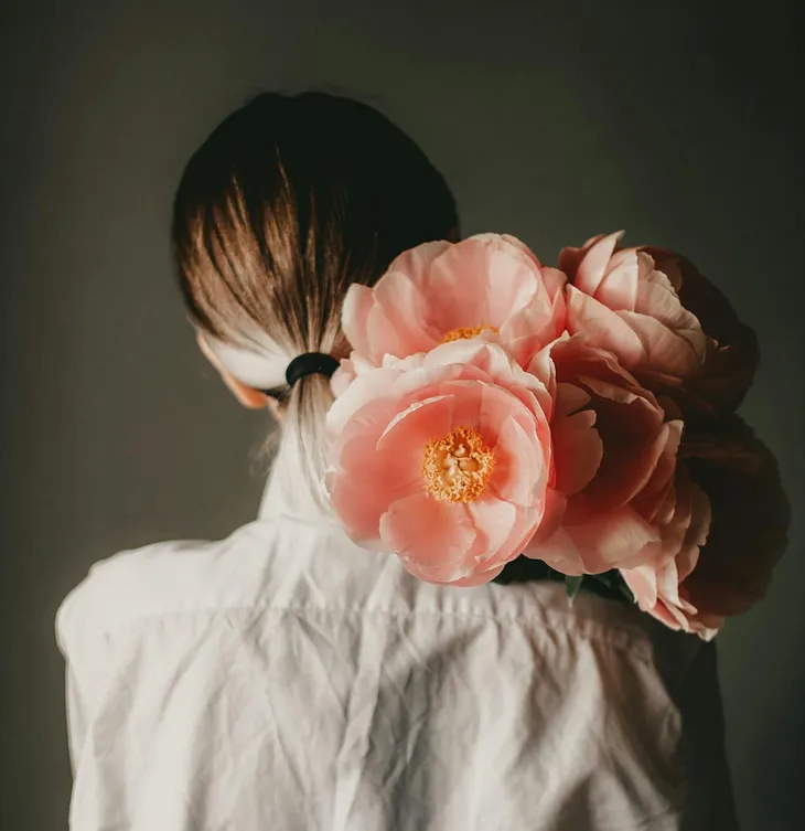 Rear view of woman in a white shirt holding pink peonies over her shoulder