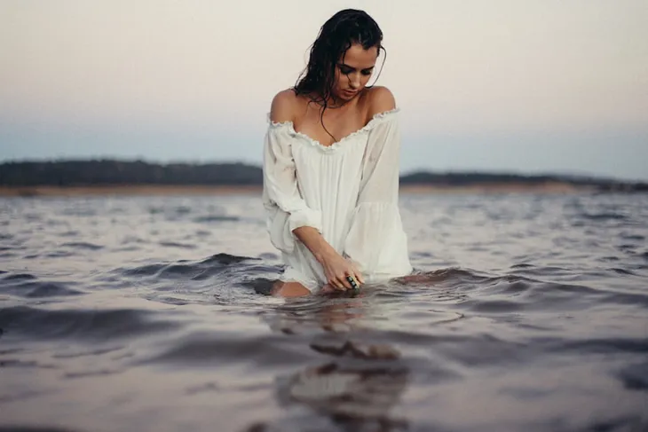 The image shows a young woman kneeling in calm water at dusk, her wet hair clinging to her face. She wears an off-the-shoulder white dress, slightly transparent from the water, giving a soft, ethereal look. Her hands rest in the water, rippling its surface, while her gaze is lowered. The background is a blurred shoreline and a gradient sky transitioning from light blue to soft pink.