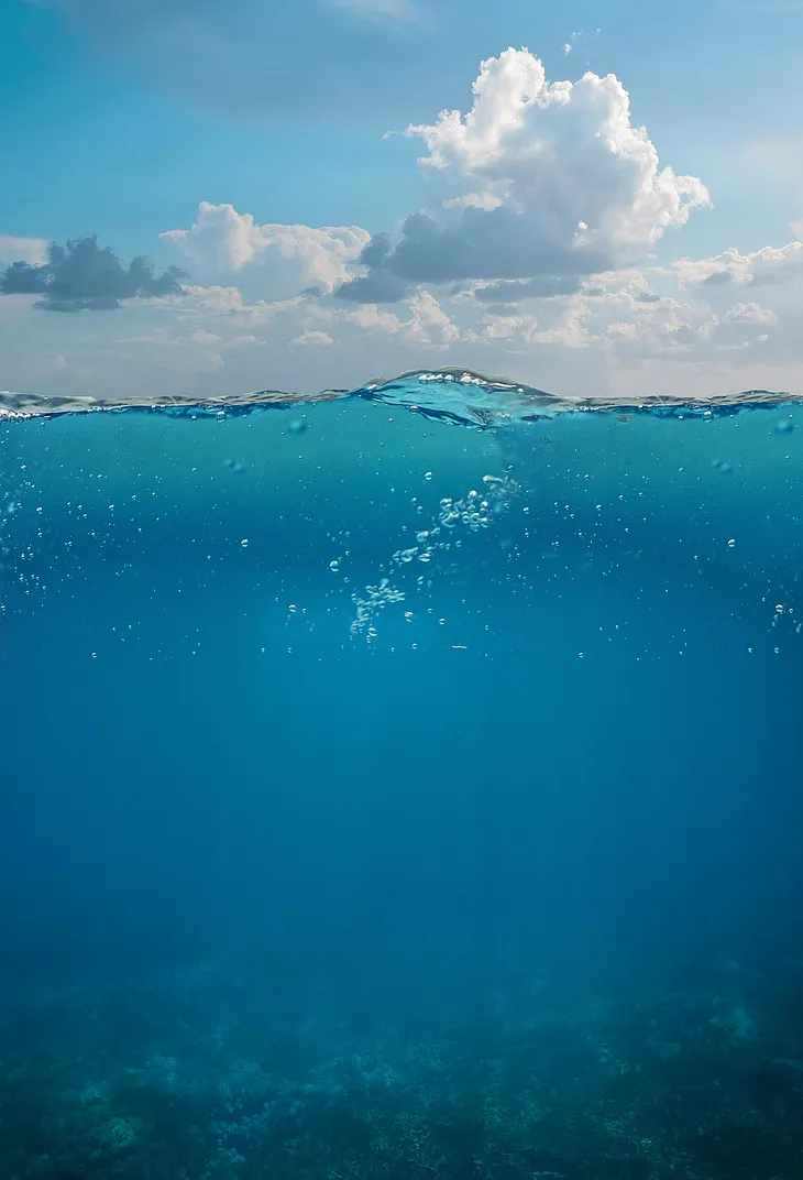 Cross-sectional view of cloudy, bright blue skies above turquoise blue waters with light ripples and air bubbles dotting across the water surface and the sea bed visible underneath.