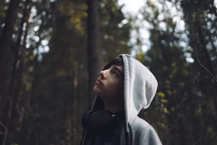 Teen in a gray hoodie gazing upward in a wooded area, looking thoughtful and introspective.