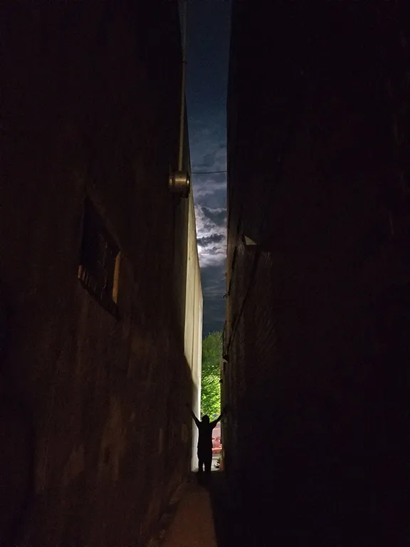 Figure stands silhouetted in the darkness between two walls, facing a mural in the distance.