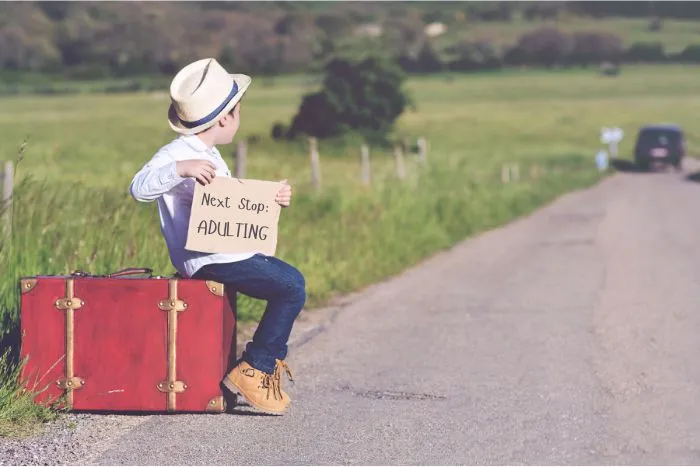 Picture of a young boy sitting on a suitcase on the side of a road holding a sign that says, “Next Stop: ADULTING.”