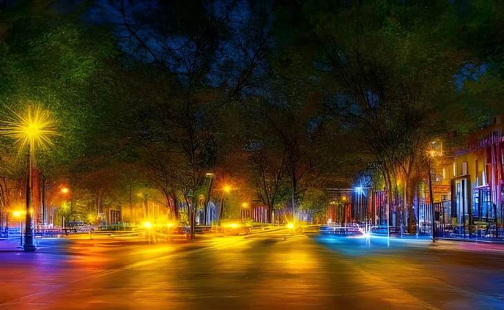 A nighttime scene of a large plaza with bright amber and blue lights here and there. Storefronts are seen on the right, also with bright lights. A magical scene.