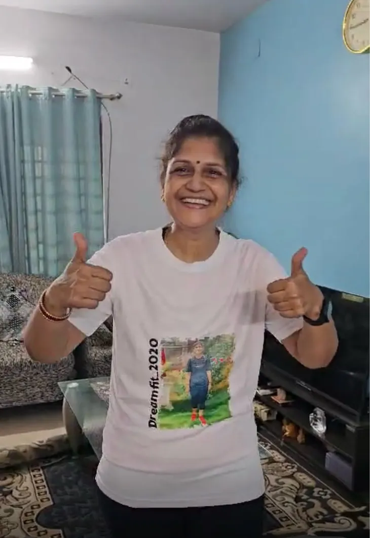 A woman smiling and giving two thumbs up, wearing a white T-shirt with the words “Dreamfit 2020” printed vertically along with an image of a person on the front. She is standing in a living room with blue curtains, a couch, and a television visible in the background. The atmosphere is casual and joyful.