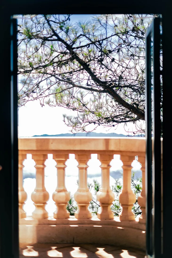 a serene view from an open doorway, framed by a balcony railing. Through the doorway, you can see a lush tree with delicate branches and a bright sky in the background. The balcony features elegant stone balusters, creating a peaceful and inviting atmosphere