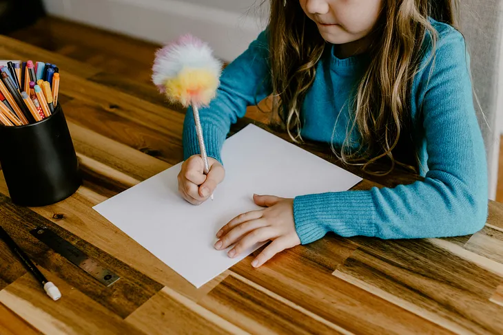 The image shows a young child in a blue sweater sitting at a wooden table, holding a colorful fluffy pen and writing on a blank piece of paper. On the table, there is a cup full of colored pencils, a ruler, and an eraser, suggesting a creative or educational activity.