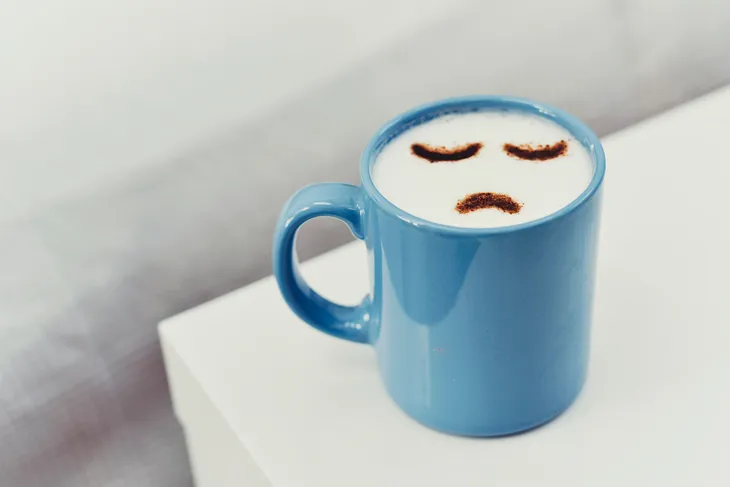 Mug of cappuccino with a frowning face representing the negative effects of caffeine and caffeine withdrawal
