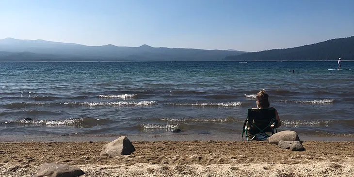Girl sitting in a low profile camp chair along the shoreline of a lake with mountains in the distance