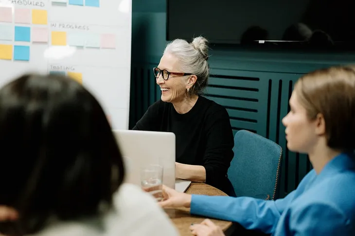 an older woman sits at a round table with a white board behind her, smiling in a direction off camera after transitioning from sex educator to sex coach.