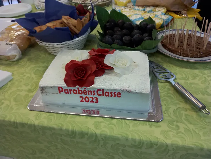 Buffet table with food. A white frosted cake with red roses that says “Parabens Classe 2023”