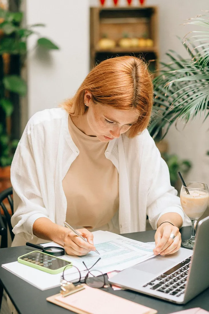 A young woman sitting at a desk with an open laptop, writing on a form.