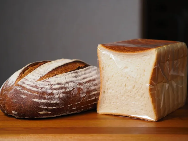 A close-up photograph shows two contrasting loaves of bread side by side on a smooth wooden surface. On the left, a round artisanal sourdough bread with a golden-brown crust displays an intricate pattern of scoring marks and a dusting of flour. On the right, a rectangular factory-produced white bread loaf wrapped in plastic is sliced into uniform pieces, showing soft, dense crumb structure. Both loaves are illuminated by soft directional lighting from above, emphasizing the textural differences
