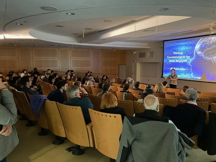 A woman speaks to a packed room of people. A large screen behind her displays a blue slide with the words, “Starting foundational AI for brain research.”