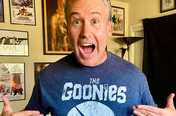 The author stand with his mouth open in happiness, pointing at his The Goonies T-shirt