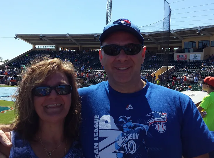 A man and woman in Bluejay shirts smile to the camera, at a spring training baseball game