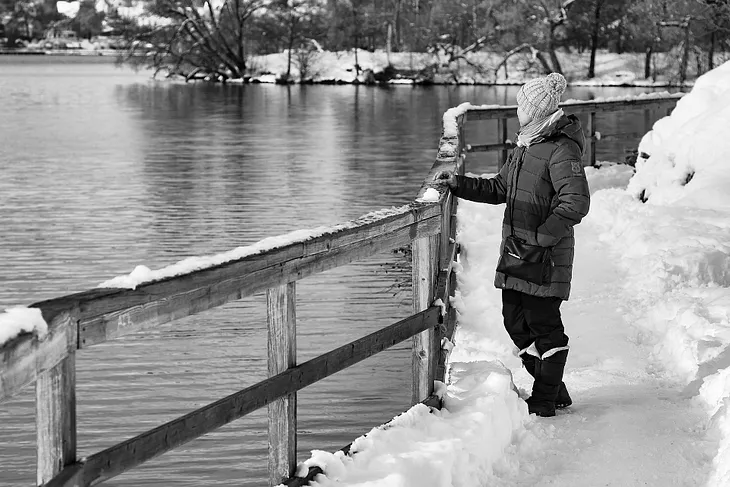 Black and white picture; woman standing in snowy landscape, overlooking a lake.