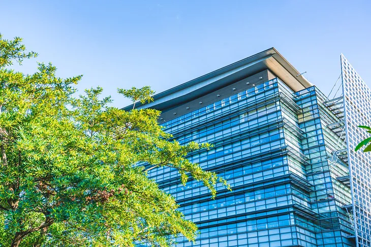 A modern glass office building reflecting the clear blue sky, with large green trees in the foreground, adding a natural contrast to the sleek architecture. The building’s angular design and clean lines are highlighted against the vibrant greenery.