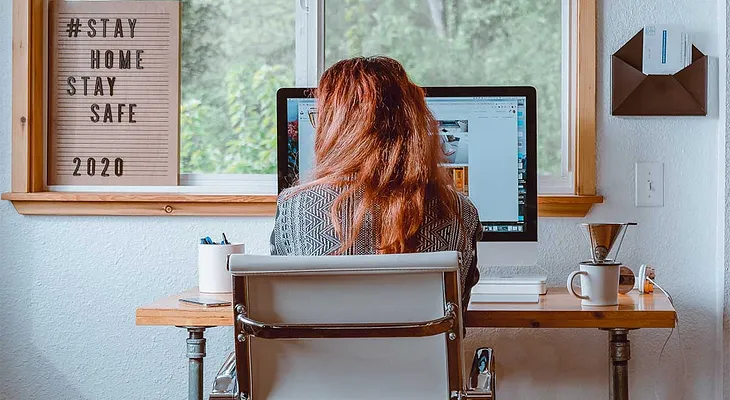 woman at her desk at home on computer. Covid19 message on wall