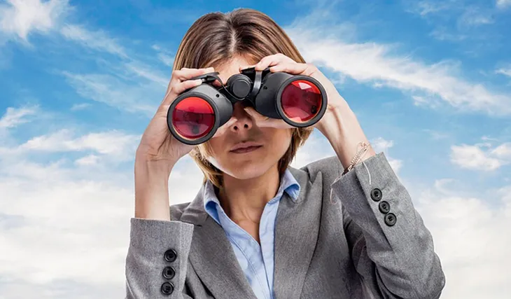 A photo of a woman looking through a pair of binoculars at the reader.