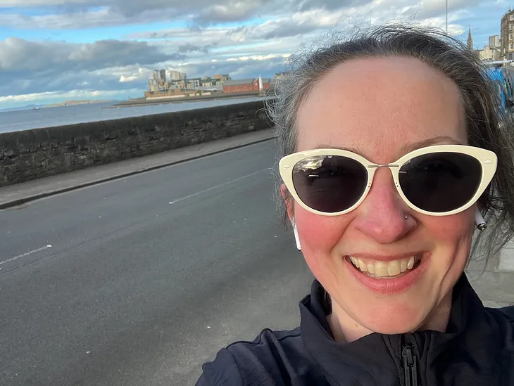 A sweaty woman with pink cheeks and white-framed sunglasses smiles at the camera. Behind her is a sea view and a road. She looks tired but happy.