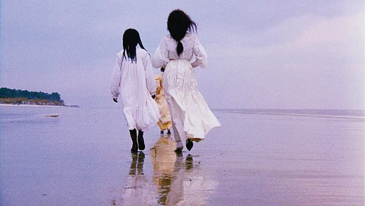 Three women dressed in white and cream dresses walking along a beach.