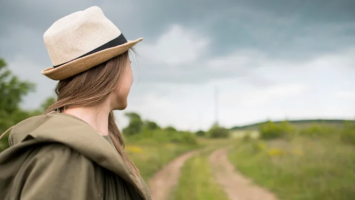 Stylish traveller with hat looking away at the dirt road behind her.