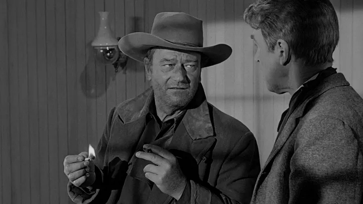 Screenshot of John Wayne and James Stewart in “The Man Who Shot Liberty Valance.” Wayne is holding a lit match.