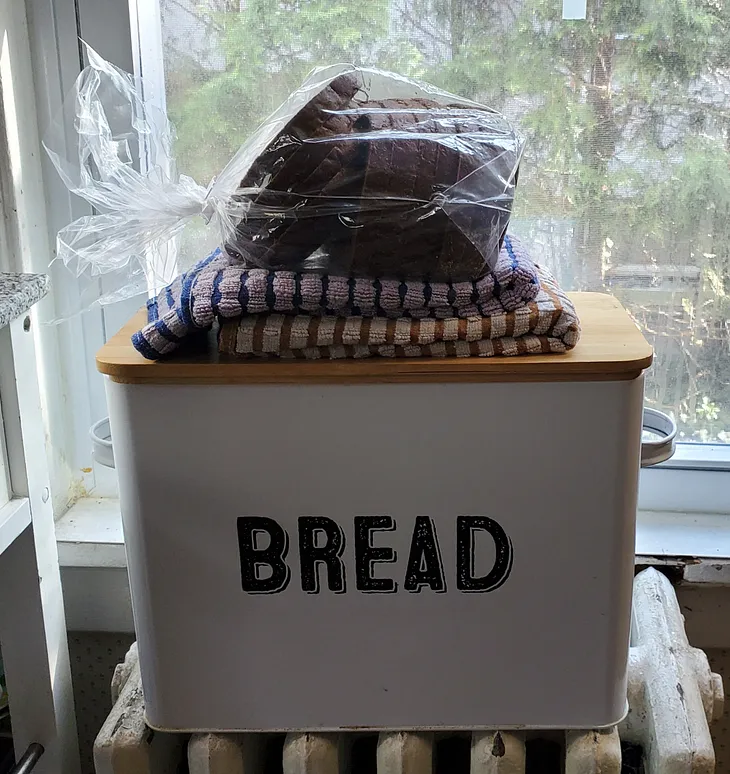 A bin that says ‘BREAD’ with towels and a loaf of bread on top of it in front of a kitchen window.