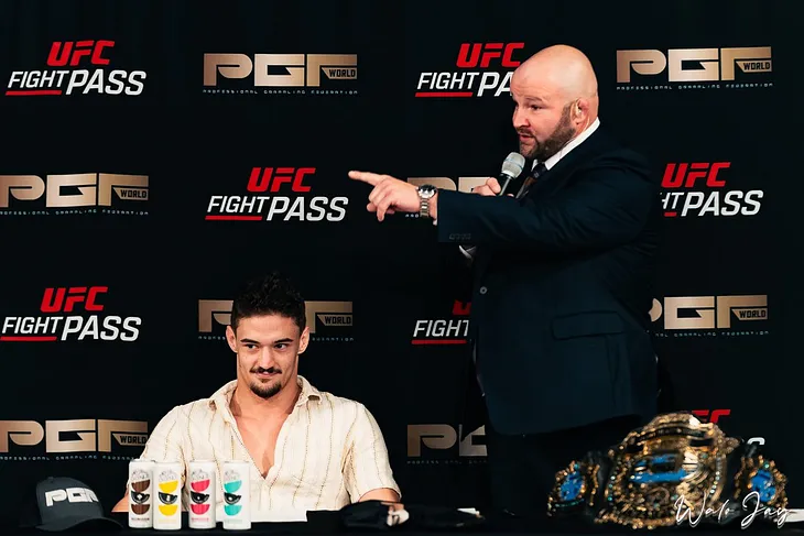 A man sits at a table with an announcer holding a microphone standing beside him and pointing.