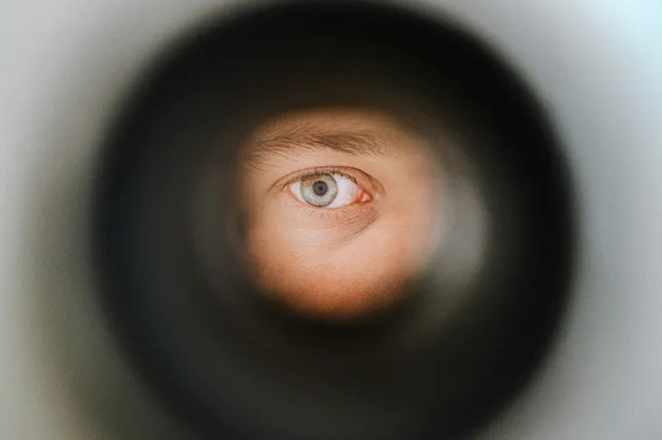 A man’s eye gazing through a peephole in a door. Only the eyebrow, one hazel eye, and the flesh just below the eye are visible.