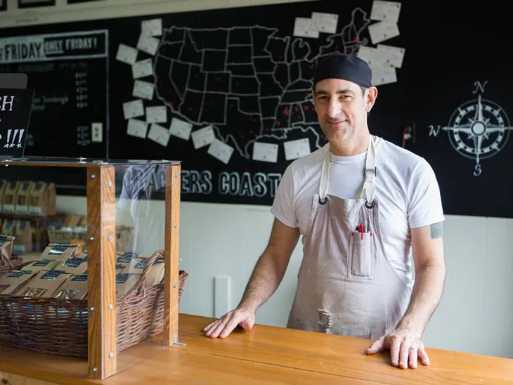 A man in an apron stands at a counter in front of a hand drawn map of the United States surrounded by note cards.