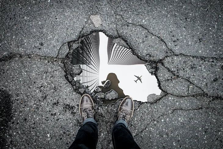 Two feet at edge of puddle reflecting an airplane in the sky
