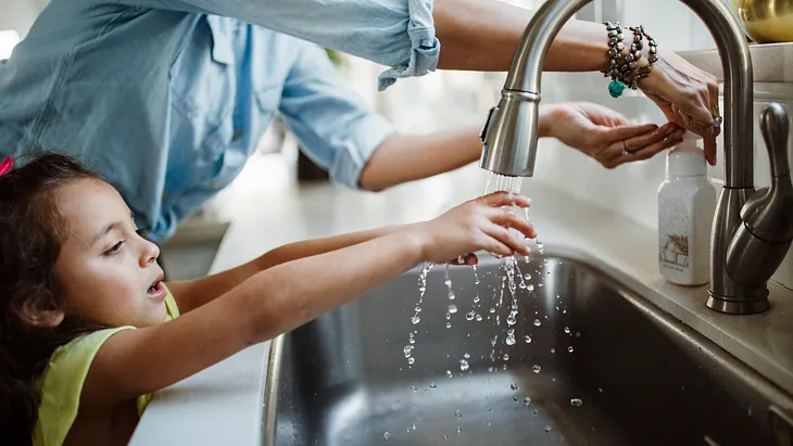 A young girl reaches up to wash her hands at a kitchen sink, assisted by an adult standing behind her. Water flows from the faucet as both their hands are under the stream, highlighting a moment of daily routine and care