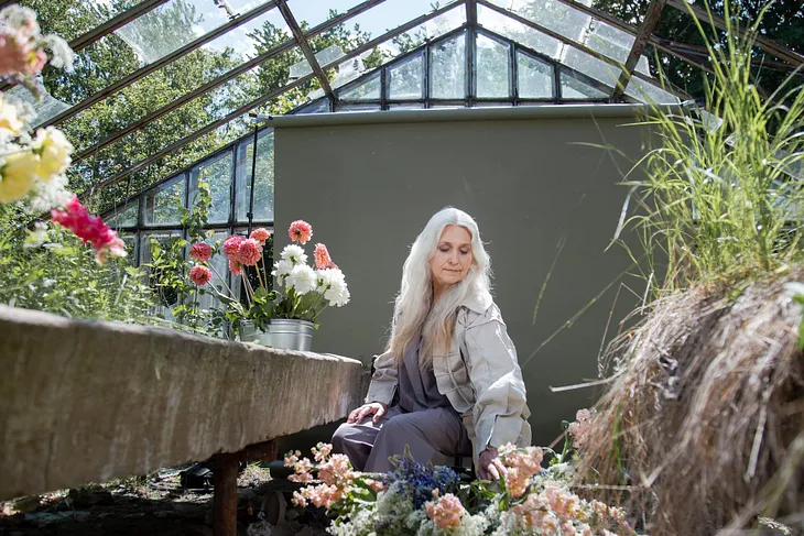 An older woman with long gray hair cutting pink, white, and red flowers in a greenhouse.
