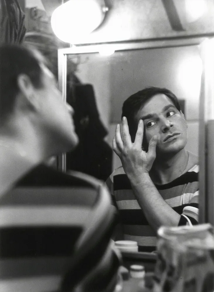 A haunting black and white photograph: a young man, striped shirt, lost in his bathroom mirror. His touch, gentle on his face, reflects a contemplative soul. The blurred edges, grainy texture, and shadows create an unsettling atmosphere.