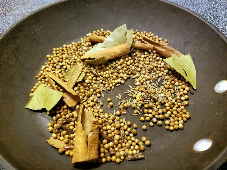 A variety of whole spices, such as bay leaves, cinnamon sticks, coriander seeds, and cumin seeds, is spread out on a black pan. The mixture suggests a preparation for roasting or toasting the spices to enhance their flavors before use in a recipe.