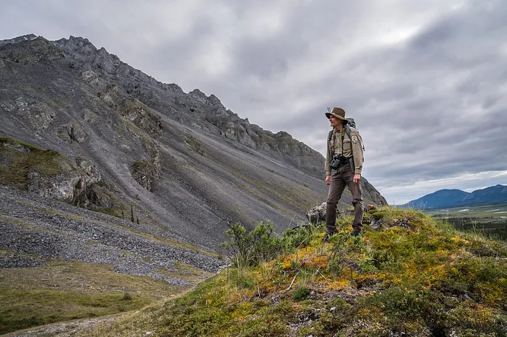 A uniformed biologist stands on a mossy hill with mountains in the background.