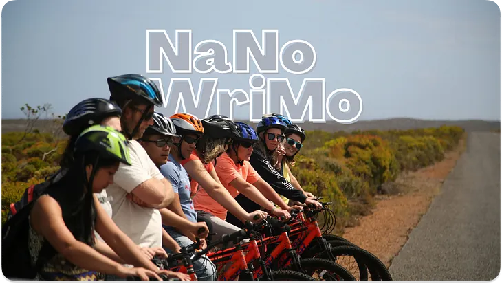 A group of bicyclists on the left side of the road. All lined up, with helmets on. In the background is NANOWRIMO in a white outline.