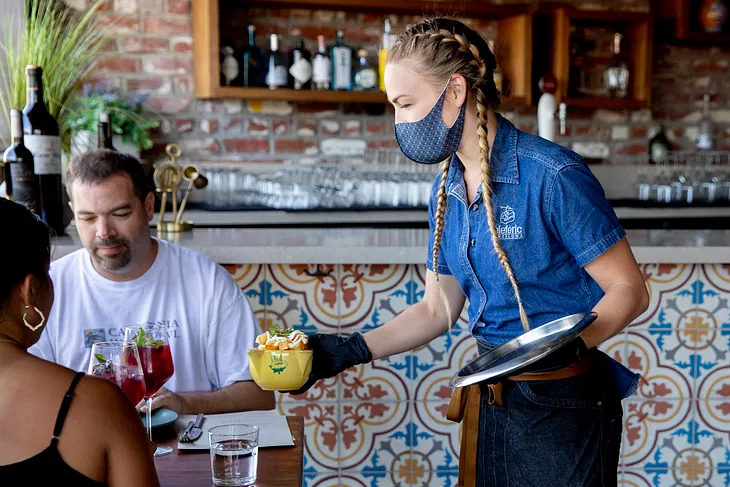 A man and a woman are served at a restaurant. The waitress is wearing a mask, they are not.