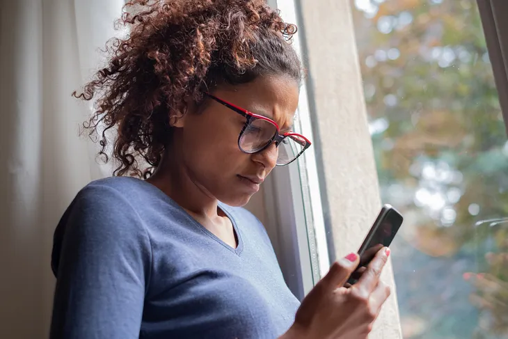 A photo of a black woman looking at her phone, looking upset.