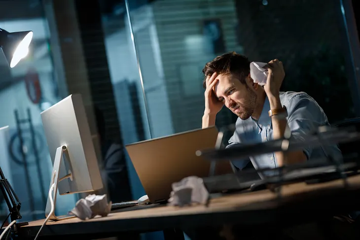 Casual man having problems at work and crumpling papers being uninspired and watching at laptop.