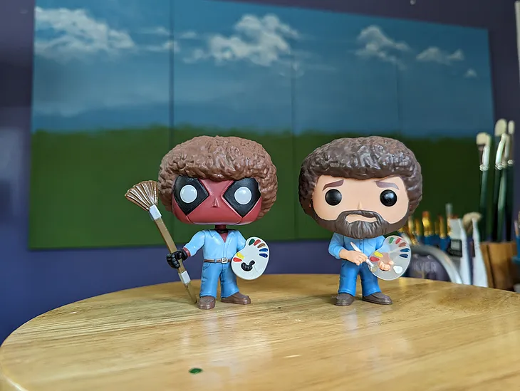 Funko Pop Bob Ross and Deadpool Bob Ross figurines placed in an art studio in front of a large 4 panel painting as inspiration for the artist herself. Which is me. Kathy LaFollett. Kathy LaFollett Murals and Canvas