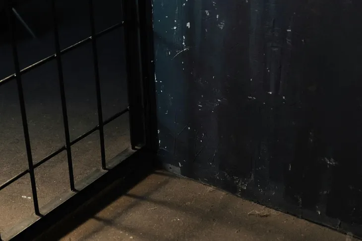 A close-up of the corner of a prison cell, with a dark wall visible on one side, and the iron bars on the other.