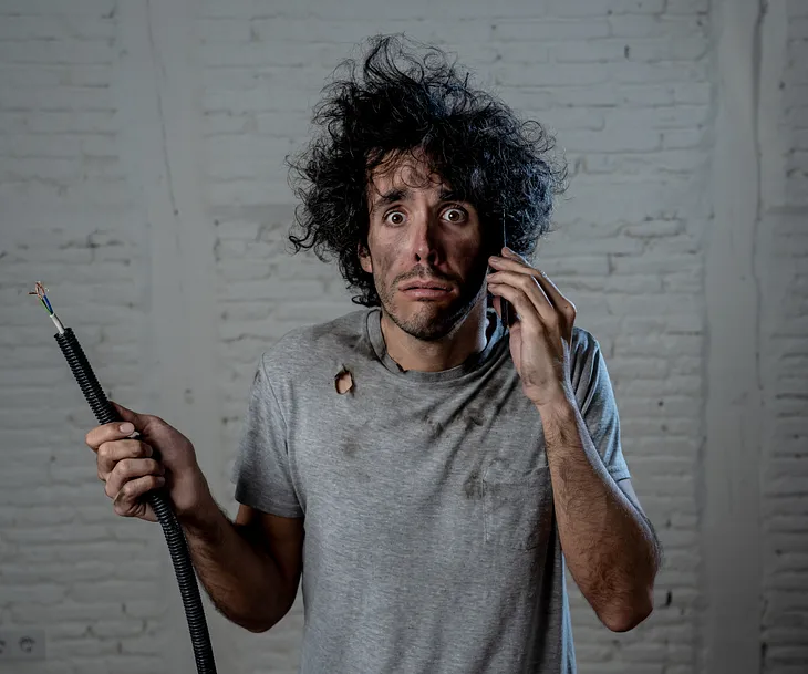 Portrait of young man holding electrical cable after domestic accident with dirty burnt funny face expression calling desperate with mobile phone asking for help. Electricity repairs and DIY concept.