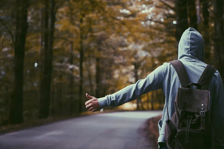 A young man stands on a roadside, hitching.