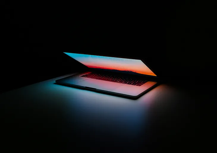 An Apple laptop open half way displaying an image of a multi-colored sky filled with pinks, blues, purples, and oranges set against a dark background.