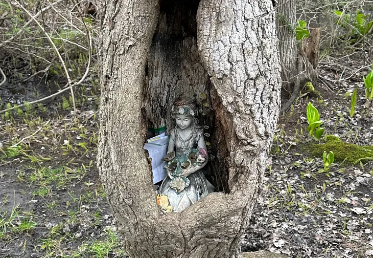 A figurine of a girl with flowers kneels in a hole within a tree on a hiking trail.