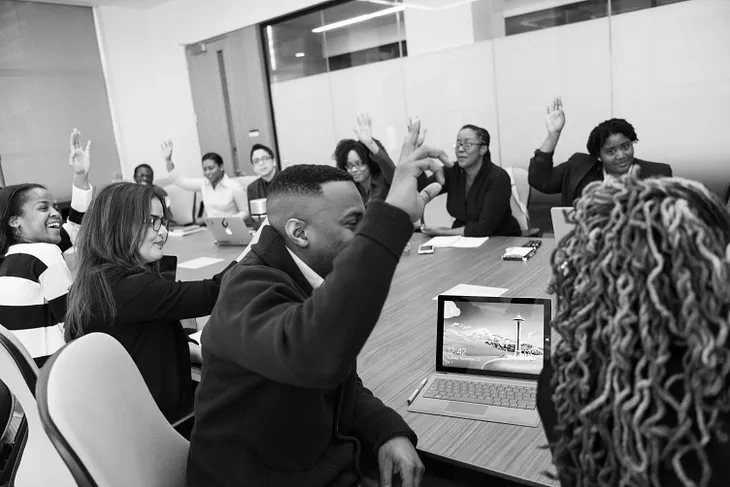 A black and white picture of several people sitting around a conference table, many of them have their hands raised. They are a variety of genders and races.