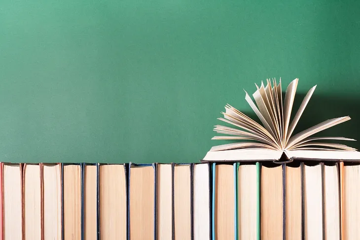 An open book sitting ontop of books lined on a shelf in front of a green wall