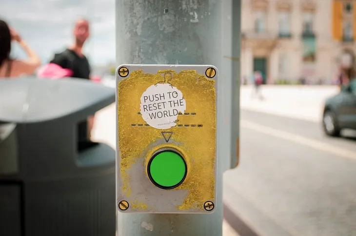 A pole in the street, with a button and the words, Push to reset the world above